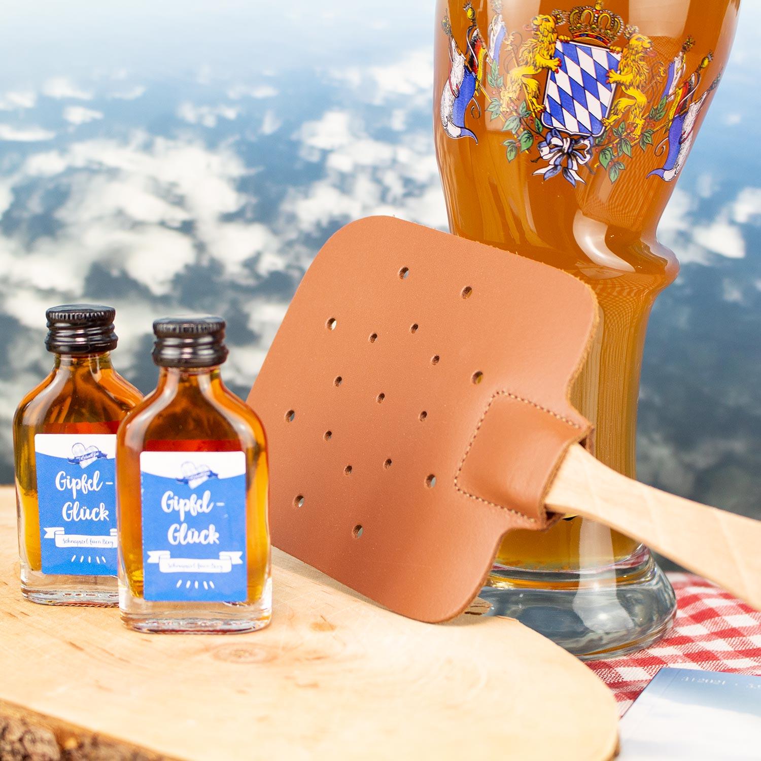 Bayerische Geschenkideen: Zwei Gipel-Glück Schnapsflaschen, Lederfliegenklatsche und Bierglas vor Alpenkulisse.