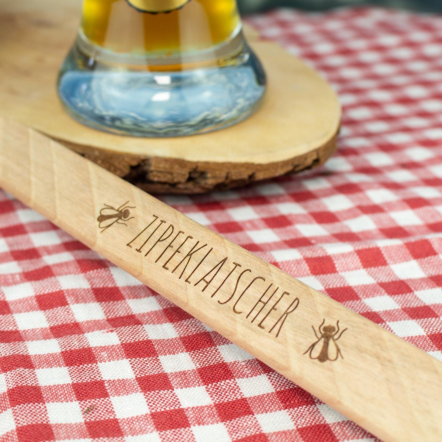 Holzlöffel Zipfeklatscher mit Gravur vor rot-weiß karierter Tischdecke und Bierglas, traditionelles Küchenaccessoire.