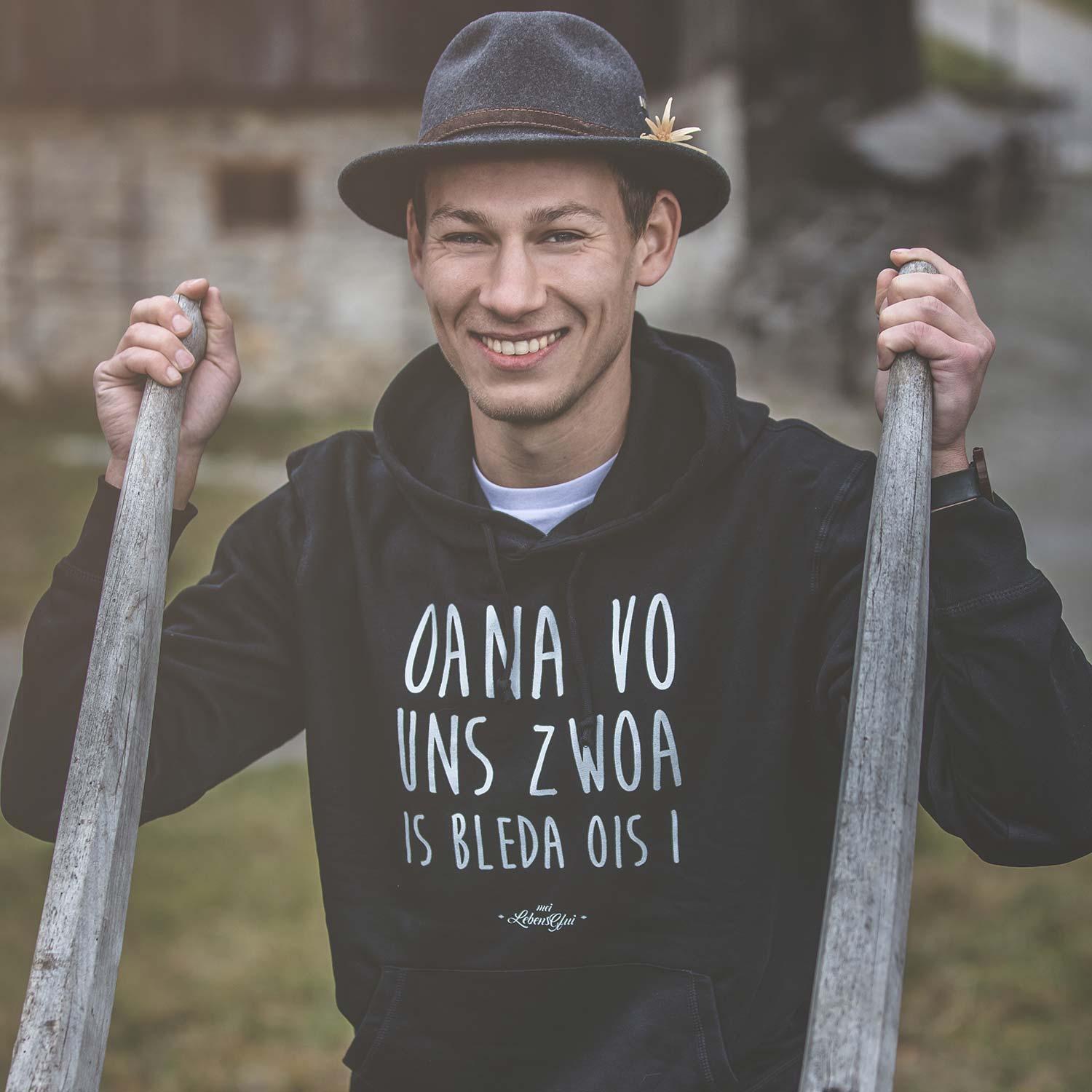 Lächelnder Mann im Hoodie Oana vo uns zwoa... und Trachtenhut, Hände auf rustikalen Holzstangen.