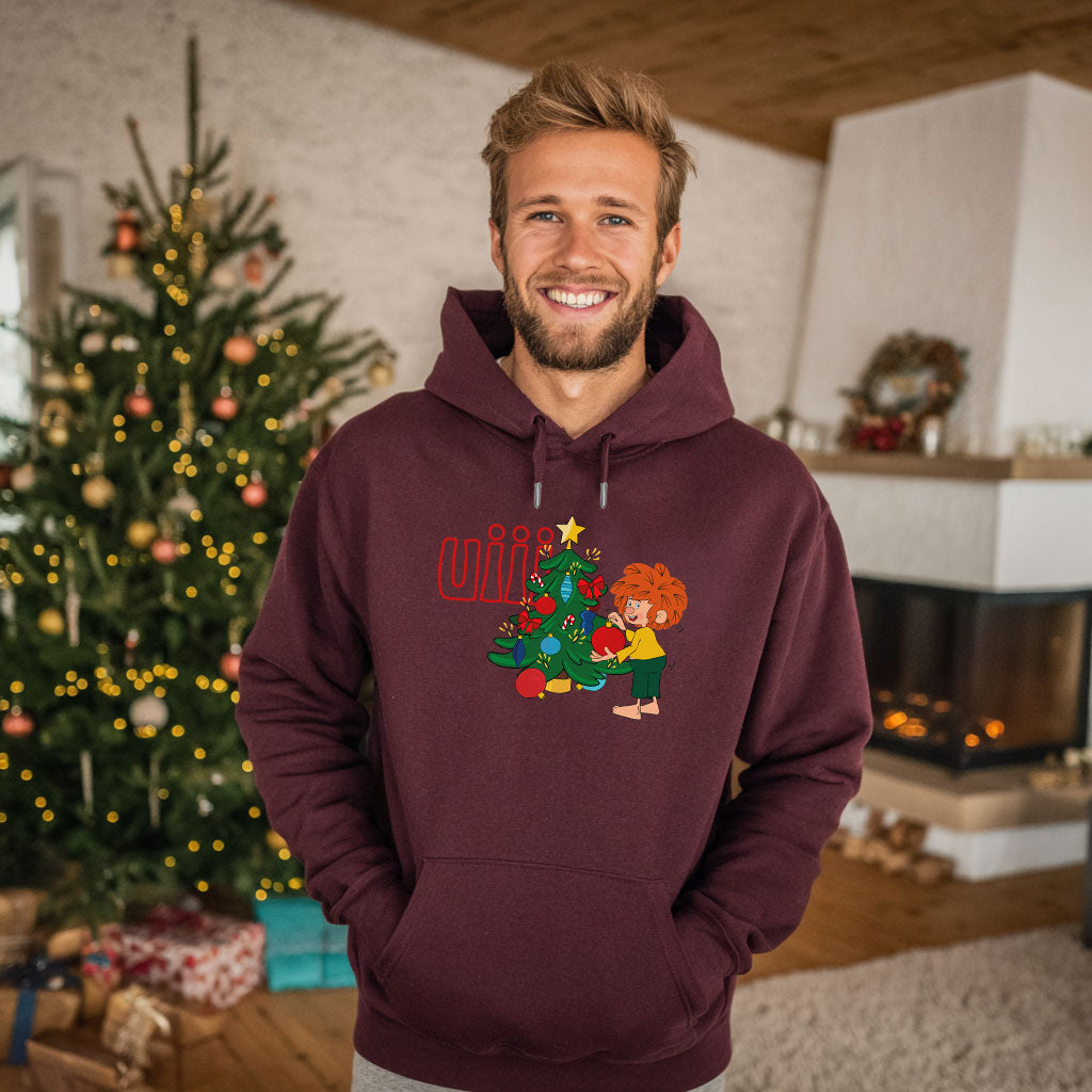 Mann trägt Pumuckl Hoodie „uiii Weihnachten“ mit Weihnachtsmotiv, während er lächelnd vor einem Tannenbaum steht.