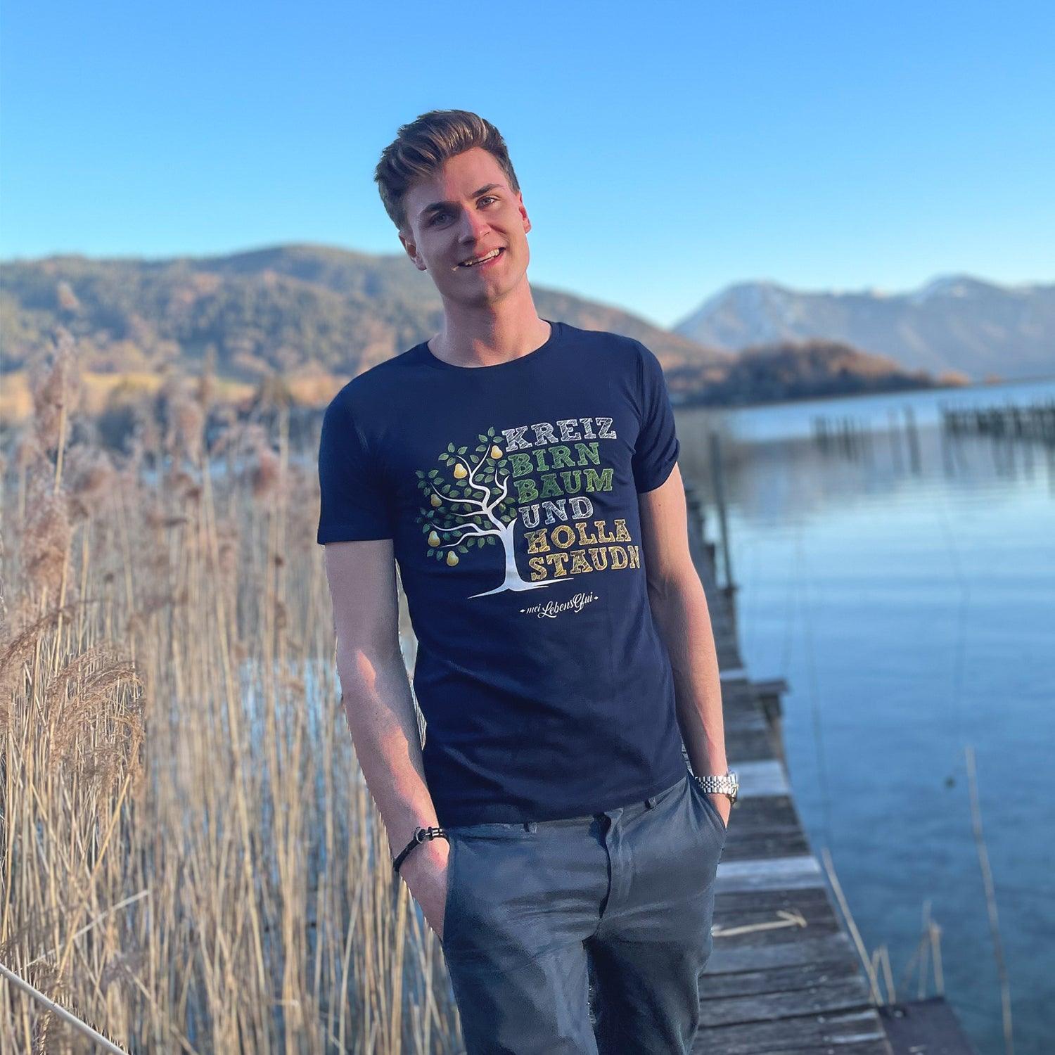 Junger Mann im marineblauen Herren T-Shirt Kreizbirnbaum mit Bergblick an einem See auf einem Holzsteg.