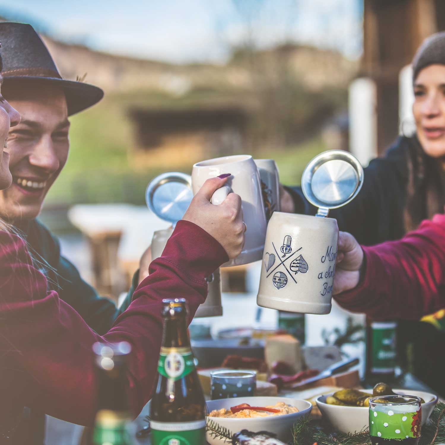 Freunde stoßen mit traditionellen Bierkrügen an einem Tisch im Freien an, dekoriert mit herzhaften Speisen wie Käse und Wurst.