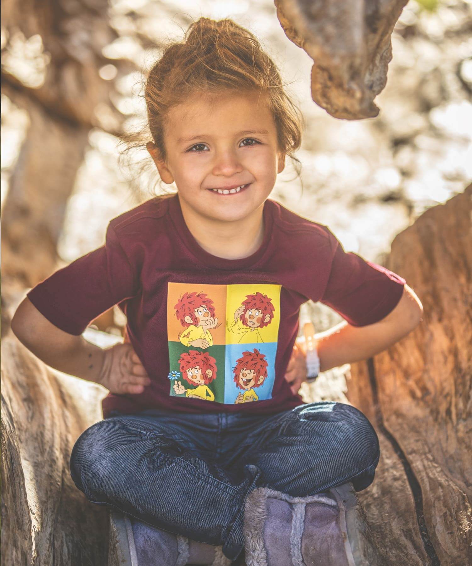 Fröhliches Kind mit Zöpfen und bordeauxrotem T-Shirt mit farbigen Kinder-Comic-Aufdruck in einem Baumstamm sitzend.
