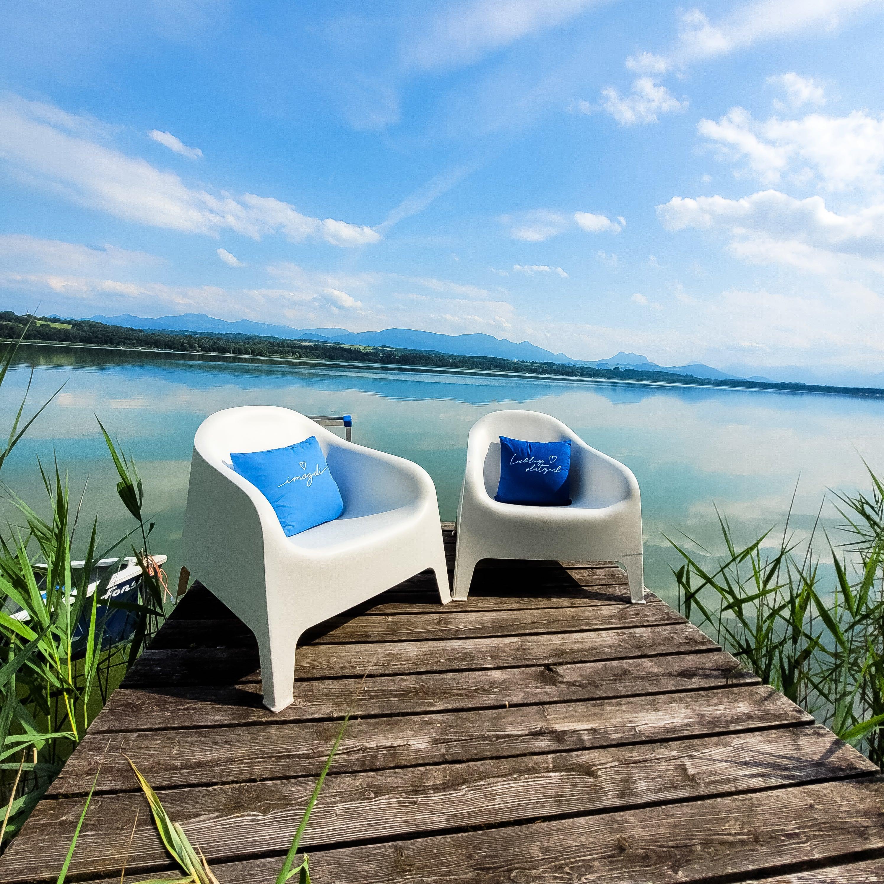 Zwei blaue Kissen "I mog di" auf weißen Stühlen auf einem Holzsteg am blauen See.