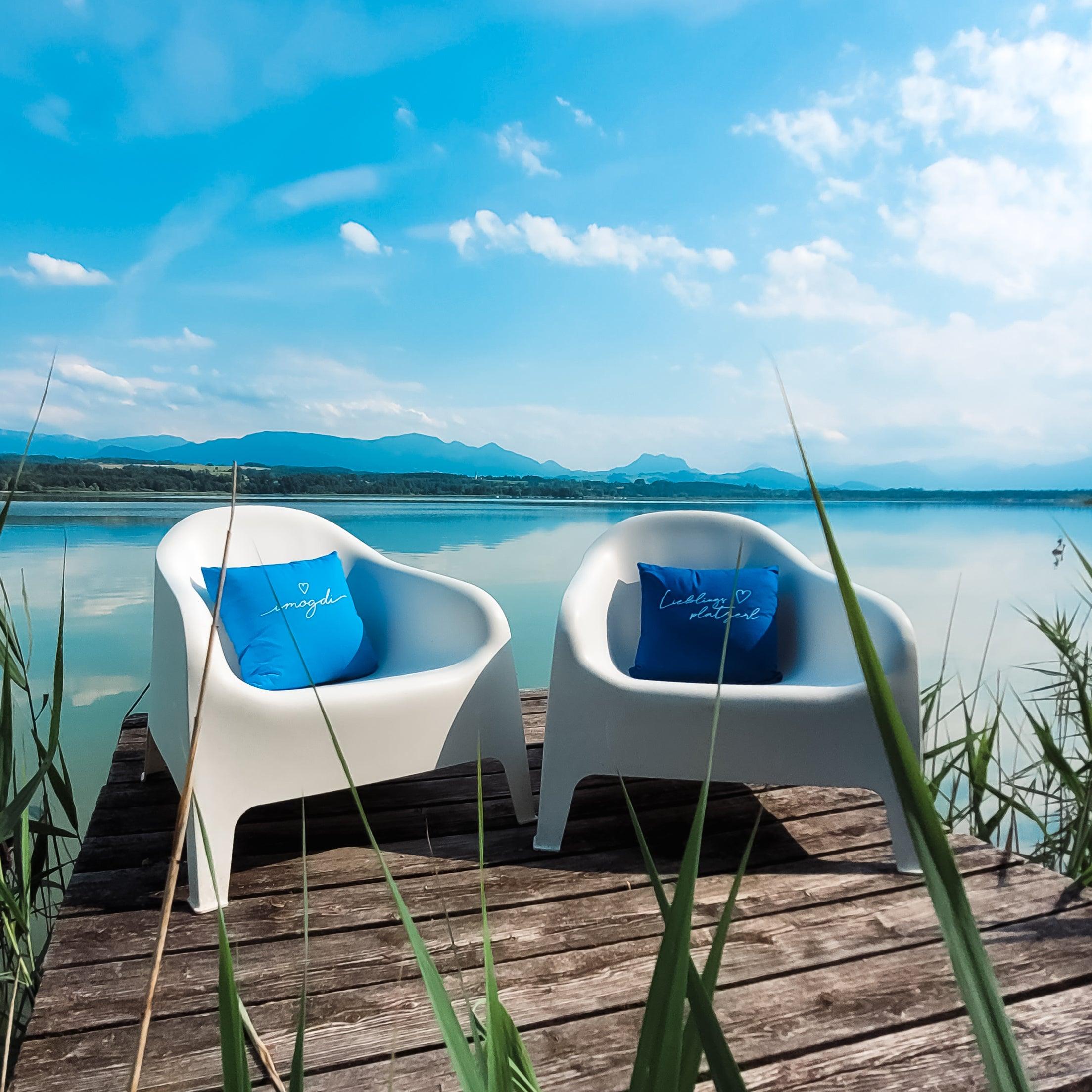 Zwei weiße Sessel auf einem Holzsteg mit blauen Kissen "Lieblingsplatzerl" am Seeufer mit Bergen.
