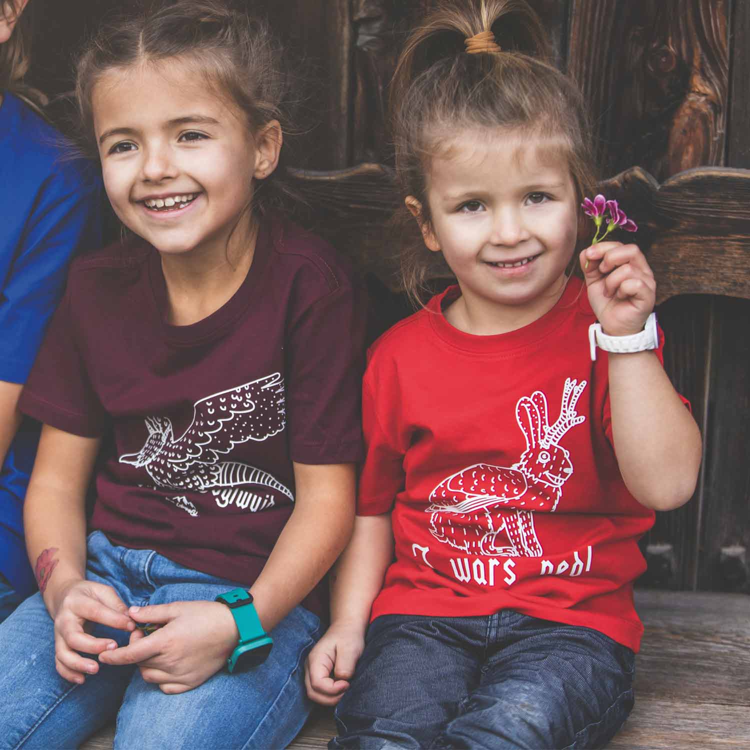 Zwei lächelnde Mädchen in Kinder-T-Shirts mit Tiermotiven und Jeans auf Holzbank.