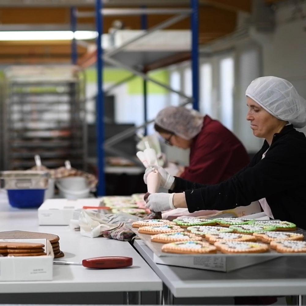 Zwei Bäckerinnen dekorieren Lebkuchenplätzchen von Hand mit Zuckerguss in einer Backstube.