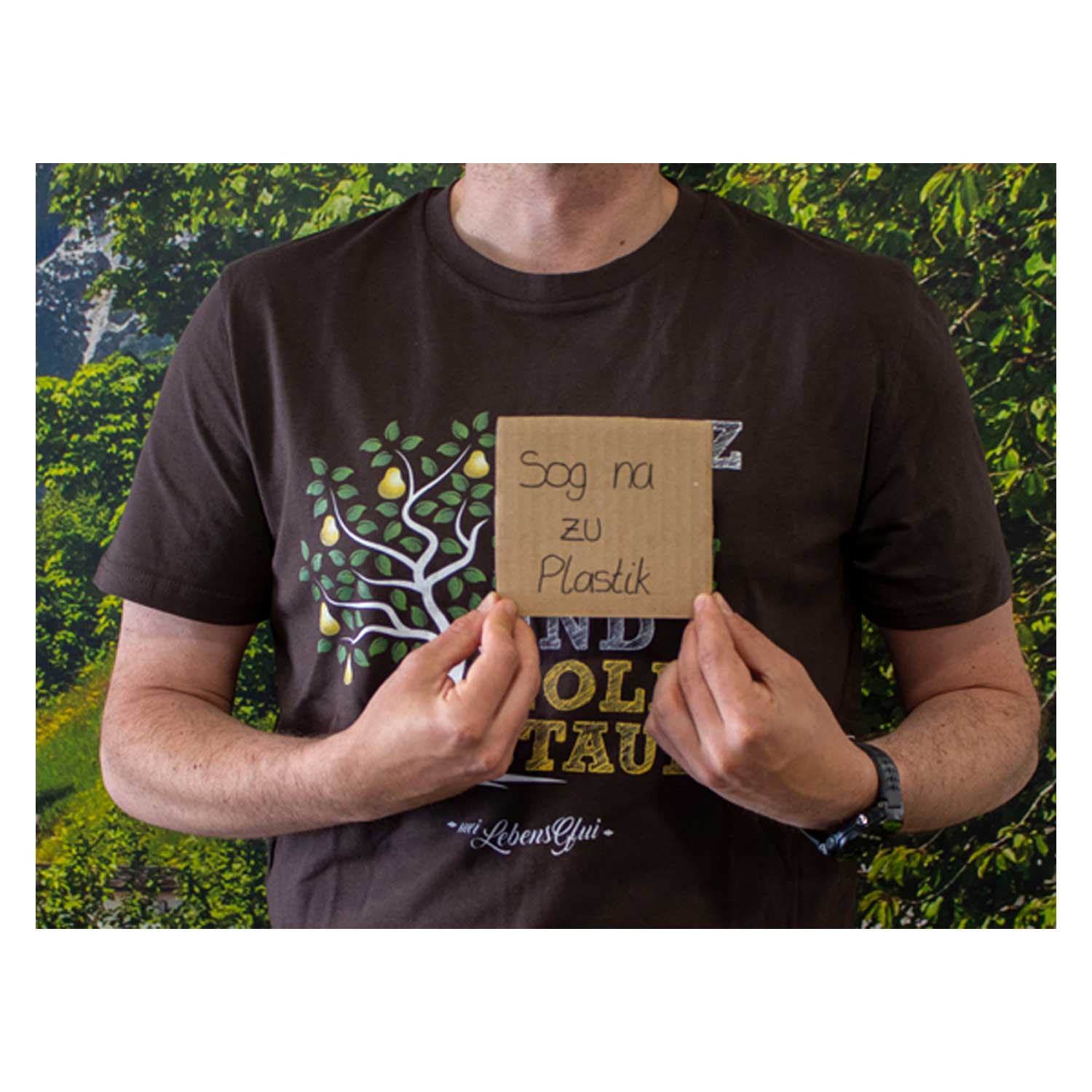 Eine Person trägt ein braunes T-Shirt mit Birnbaum-Motiv und hält ein Schild: Sog na zu Plastik