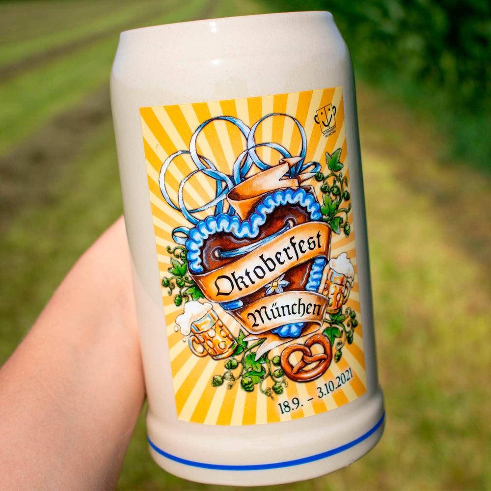 Offizieller Oktoberfestkrug 2021 mit blau-gelbem Herz-Design, Brezel, Bierkrügen und Münchenschriftzug, in Hand gehalten.