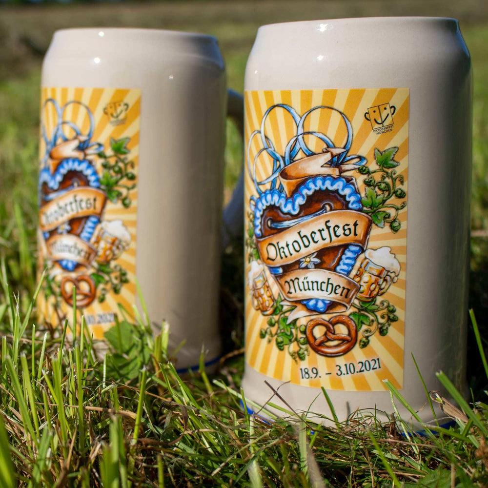 Zwei offizielle Oktoberfestkrüge 2021 mit Herzdekor und Bier-Motiven auf grünem Gras im Sonnenlicht.