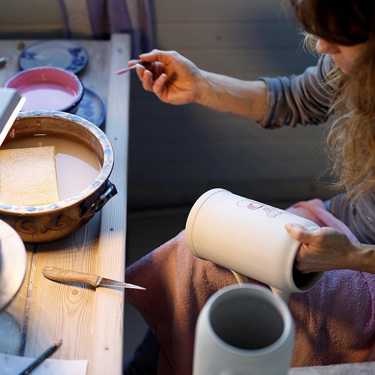 Künstlerin bemalt handgefertigten Keramik-Bierkrug mit feinem Pinsel an hölzernem Arbeitstisch in Töpferei.