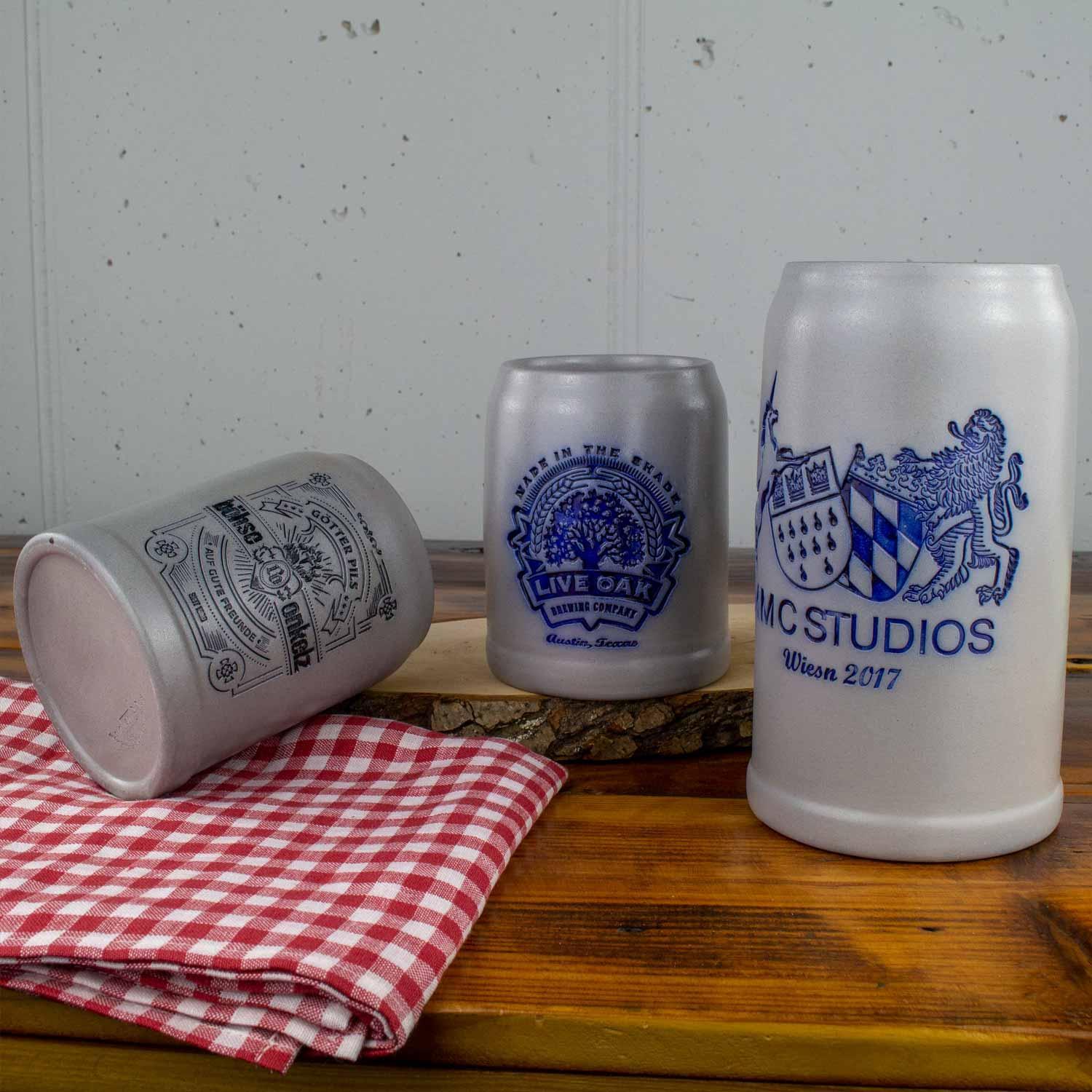 Keramik-Bierkrüge mit blauen Logos und Beschriftungen, liegend und stehend auf Holztisch mit karierter Serviette.