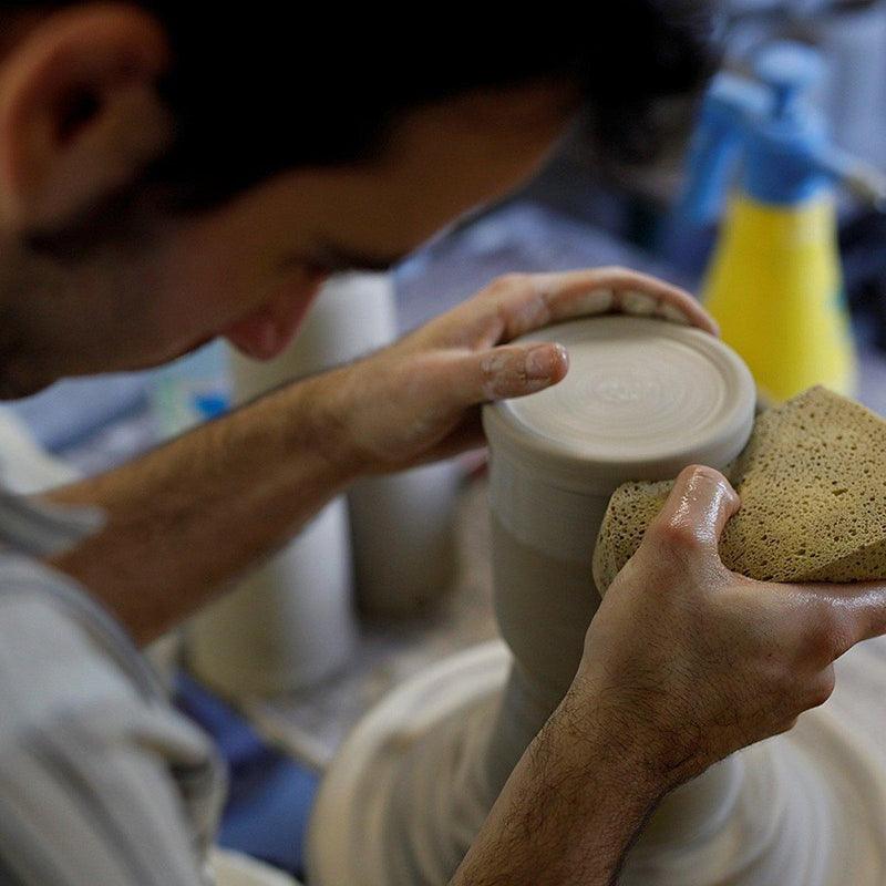 Töpfer formt einen PREMIUM Bierkrug Oberbayern aus Ton auf der Drehscheibe mit einem feuchten Schwamm und Händen.