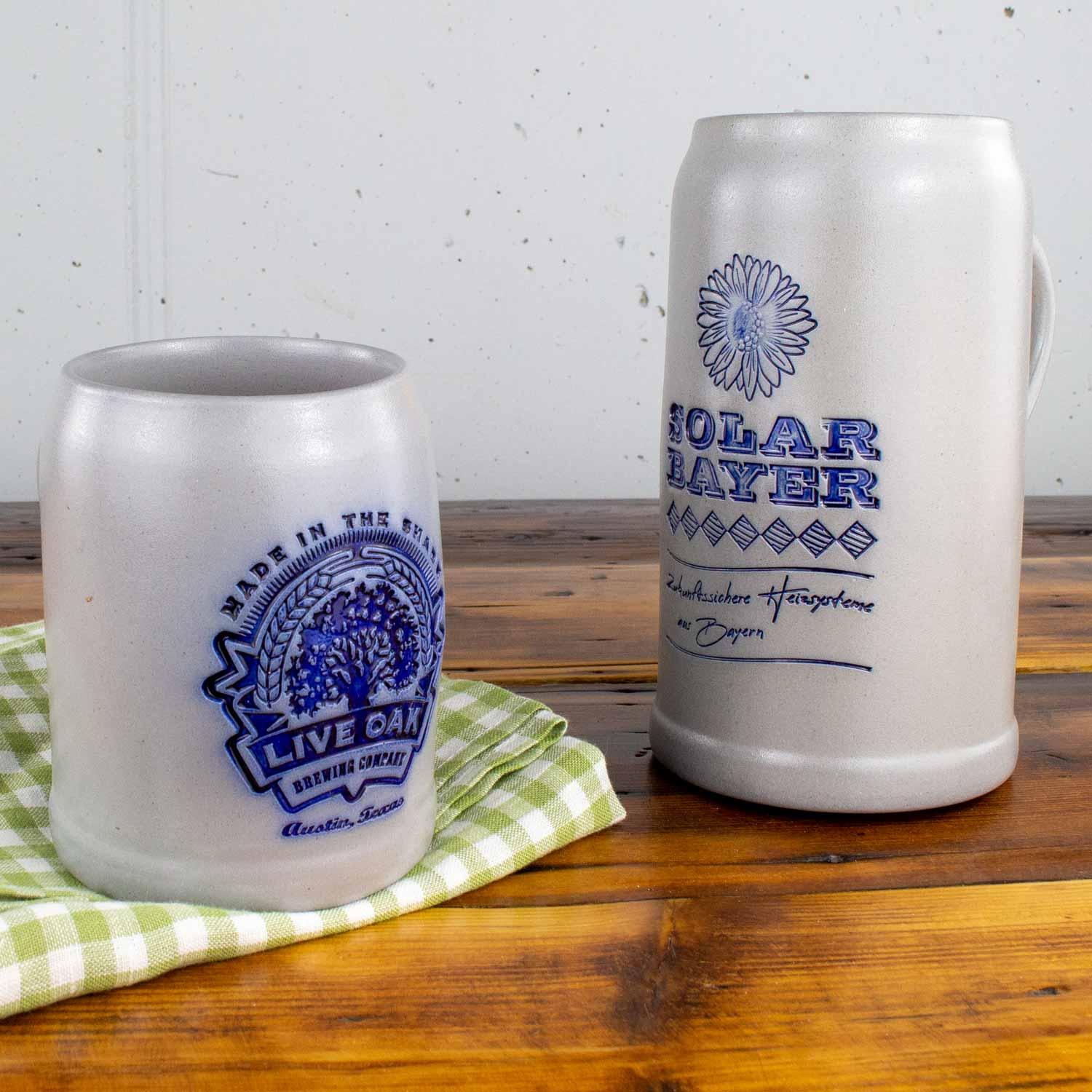 Zwei hellgraue Bierkrüge aus Keramik mit blauen Verzierungen und Logos auf rustikalem Holztisch.