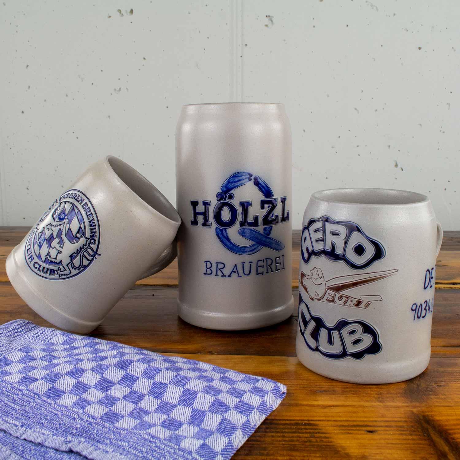 Drei individuelle Steinbierkrüge mit blauen Aufdrucken, darunter "Brauerei Hölzl", auf Holztisch mit kariertem Tuch.