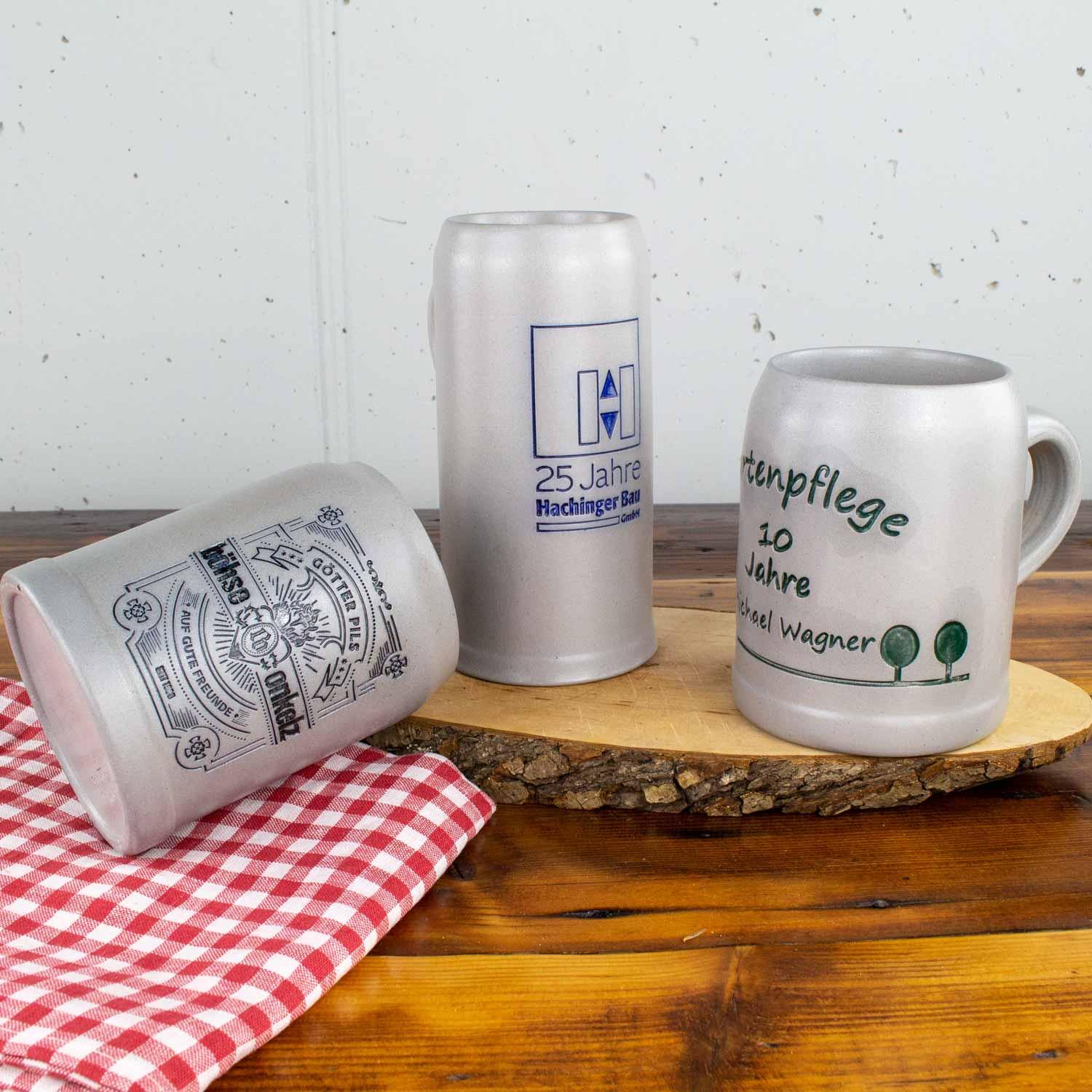 Drei verschiedene Keramik-Bierkrüge, zwei aufrecht mit Inschriften, einer liegend auf rot-weiß kariertem Tuch.
