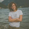 Junge Frau im weißen Damen T-Shirt "Habe die Ehre" mit verschränkten Armen vor See-Panorama.