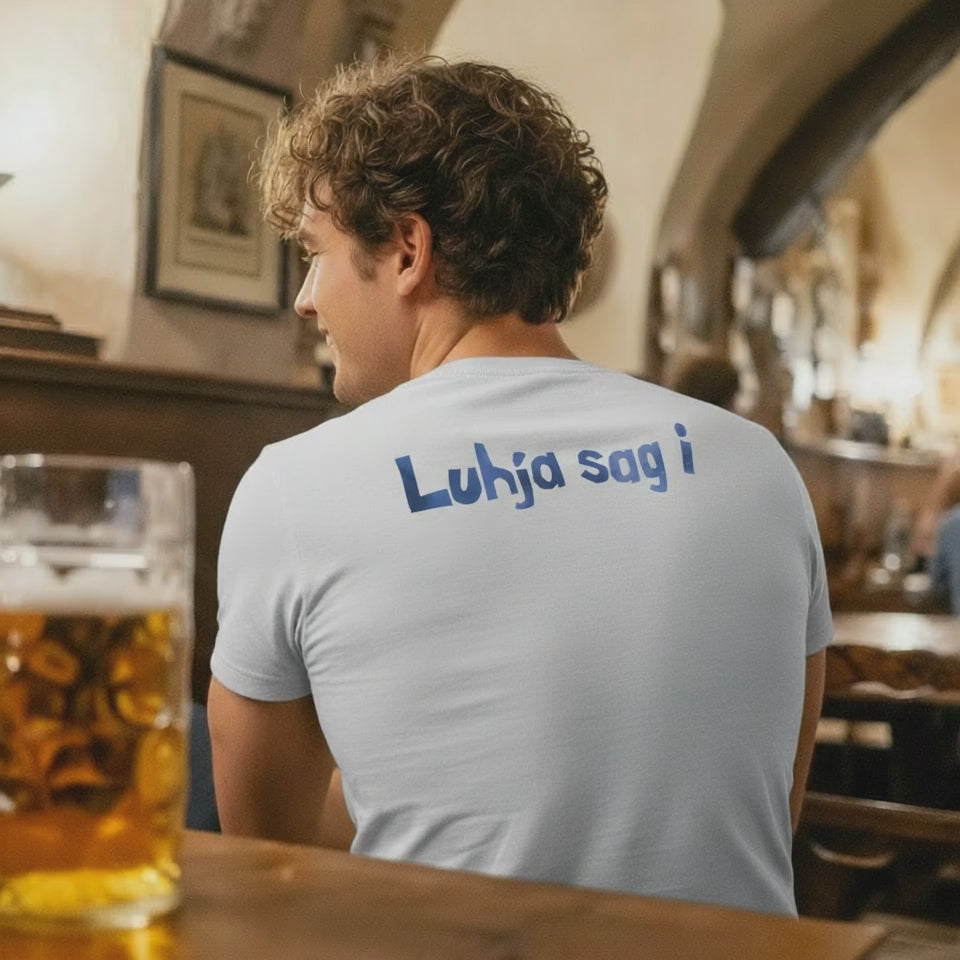 Rückenansicht eines Mannes in einem hellblauen Herren T-Shirt mit bayerischem Spruch "Luhja sag i" an einer Bar, daneben ein volles Bierglas.