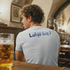 Rückenansicht eines Mannes in einem hellblauen Herren T-Shirt mit bayerischem Spruch "Luhja sag i" an einer Bar, daneben ein volles Bierglas.