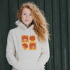 Frau mit roten Lockenhaaren trägt beigen Pumuckl Hoodie mit „Schnappschuss“-Motiv, lässig an Holzwand lehnend.