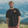 Lachender Mann in schwarzem ®Pumuckl T-Shirt "Nei da" mit Brusttasche vor alpiner Landschaft.