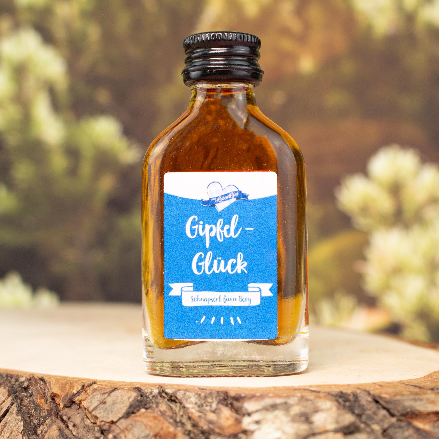 Flasche "Gipfel-Glück" Schnapserl mit blauem Etikett auf rustikaler Holzscheibe in natürlicher Umgebung.